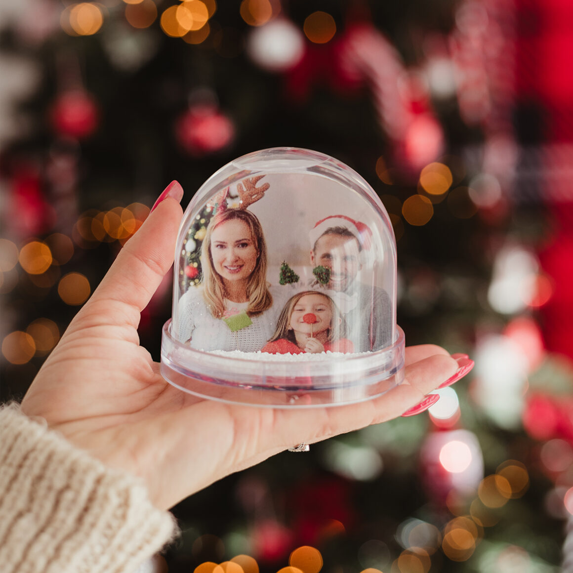 Női kézben látható egy hópihés hógömb. Benne fénykép látható egy családról. Háttérben elmosódott karácsonyi fények láthatóak